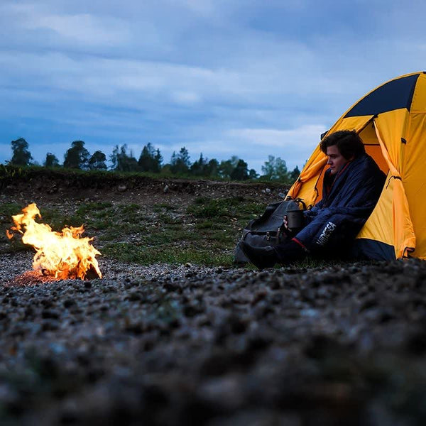 Campeur près d'une tente jaune, utilisant un sac de couchage pour se réchauffer près d'un feu de camp