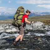 Homme traversant une rivière avec des bâtons de marche ultralégers, sac à dos vert, paysage montagneux